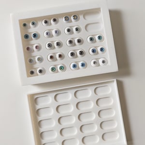 May include: A white tray filled with various sizes of glass eyes in shades of blue, brown, green, and gray. The eyes are arranged in rows within the tray. A second, empty white tray is below.