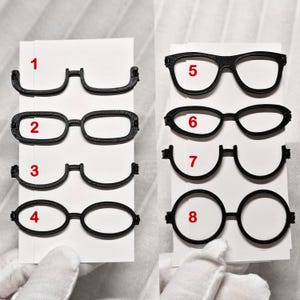 Può includere: Otto montature di occhiali neri di vari design sono mostrate su cartoncini bianchi, numerate da 1 a 8 in rosso. Le montature includono forme rettangolari, ovali e rotonde. L'immagine mostra diversi stili di occhiali.