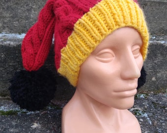May include: A red and yellow knitted hat with black pom-poms. The hat has a long, pointed top and a cable knit pattern.