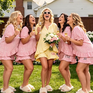 Op de afbeelding: Vijf mensen lachen in een tuin en houden champagneglazen vast. Vier dragen roze gingham pyjamasets met ruches en bijpassende slippers. Eén persoon draagt een gele jurk en houdt een boeket en witte ballonnen vast. De scène speelt zich af voor een huis.
