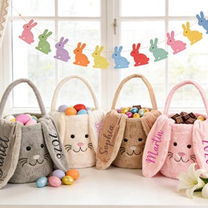 Puede incluir: Cuatro cestas de Pascua de felpa en gris, crema, beige y rosa, cada una con orejas de conejo y una cara sonriente. Las cestas están llenas de huevos y golosinas de colores. Una guirnalda de conejitos de colores cuelga en el fondo.