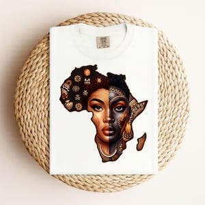 Könnte beinhalten: Weißes T-Shirt mit einer Grafik des afrikanischen Kontinents, die das Gesicht einer Frau in zwei Hälften teilt, eine hell und eine dunkel, mit aufwendigen Mustern. Das T-Shirt wird auf einer gewebten Oberfläche präsentiert.