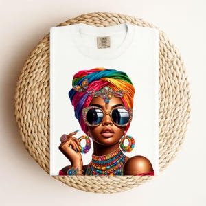 Könnte beinhalten: Weißes T-Shirt mit einer lebendigen Illustration einer Person, die einen farbenfrohen Turban, Sonnenbrille und Schmuck trägt. Das Design umfasst einen Regenbogen an Farben und komplizierte Details, wodurch ein kühnes und künstlerisches Statement entsteht. Das Shirt ist auf einer gewebten Oberfläche gefaltet.