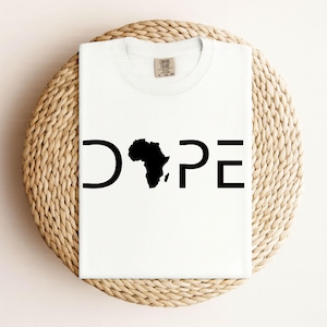 Könnte beinhalten: Weißes T-Shirt mit dem Wort "DOPE" in schwarzen Buchstaben. Das "O" ist durch eine schwarze Silhouette des afrikanischen Kontinents ersetzt. Das T-Shirt liegt auf einer runden, gewebten Unterlage.