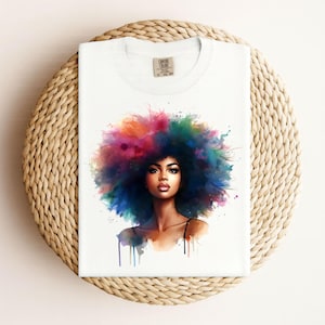 Könnte beinhalten: Weißes T-Shirt mit einer Aquarellillustration einer Frau mit bunten Haaren. Das Kunstwerk zeigt Blau-, Grün-, Rosa- und Orangetöne. Das T-Shirt ist gefaltet und auf einer gewebten, kreisförmigen Oberfläche platziert.