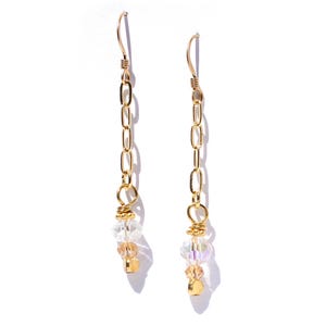 May include: A pair of gold-colored dangle earrings. Each earring features a chain design with clear and iridescent beads, and a small gold-colored square bead. The earrings have a hook closure.