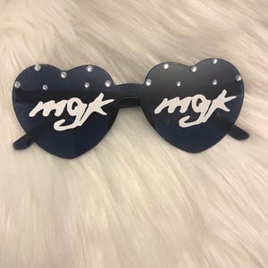 Puede incluir: Gafas de sol en forma de corazón con cristales de color azul oscuro. Las gafas tienen el texto "mgk" en blanco en cada lente y están adornadas con pequeños diamantes de imitación brillantes. La montura es negra.