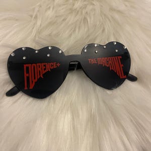 Può includere: Occhiali da sole neri a forma di cuore con la scritta rossa "FLORENCE + THE MACHINE". Gli occhiali hanno piccoli strass trasparenti lungo la parte superiore della montatura. Gli occhiali sono su una superficie bianca e soffice.