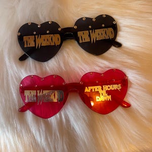 May include: Two pairs of heart-shaped sunglasses. The top pair is black with gold text reading THE WEEKND on both lenses and small rhinestones along the top rim. The bottom pair is red with gold text reading THE WEEKND and AFTER HOURS TIL DAWN.