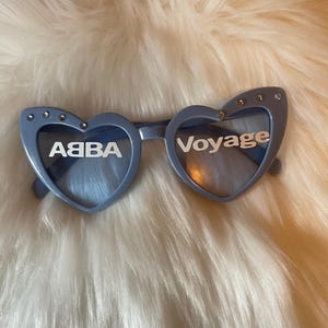 May include: Pale blue heart-shaped sunglasses with the words "ABBA" and "Voyage" in white. The frames have small silver studs. The sunglasses rest on a fluffy, white surface.