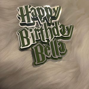 Può includere: Un cake topper a strati con le parole "Happy Birthday Bella" in una combinazione di colori verde e argento. Il testo ha un effetto 3D, con uno strato di ombra. Il topper è progettato per una festa di compleanno.