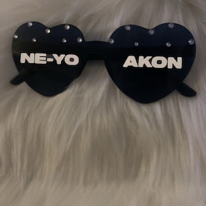 Może przedstawiać: Czarne okulary przeciwsłoneczne w kształcie serca z napisem "NE-YO" i "AKON" w białych literach. Okulary są ozdobione małymi, błyszczącymi dżetami. Tło to puszysta, biała powierzchnia.