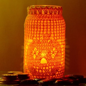 May include: A glass jar with a crocheted orange cover, lit from within, creating a warm glow. The jar is sitting on a surface with coins scattered around it.
