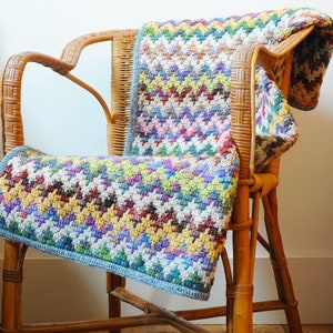 May include: A colorful crocheted throw blanket draped over a wooden chair. The blanket features a chevron pattern in shades of blue, green, yellow, pink, and white.