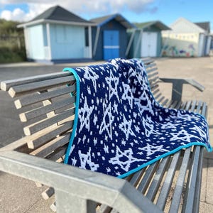 Può includere: Una coperta all'uncinetto blu scuro con un motivo a stelle bianche ripetuto e un bordo turchese brillante. Il tessuto è drappeggiato su una panchina in metallo a doghe, mettendo in risalto l'intricata lavorazione del filato e il contrasto.