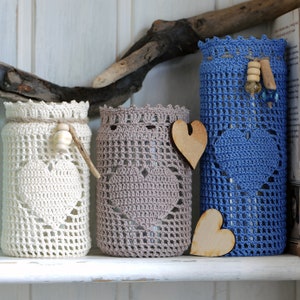 May include: Three glass jars with crocheted covers in white, gray, and blue. The jars are decorated with wooden hearts and beads.