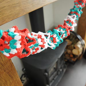 Puede incluir: Una guirnalda de crochet roja, blanca y azul verdoso con un patrón repetitivo de cuadrados. La guirnalda está colgada sobre una estufa de leña negra.