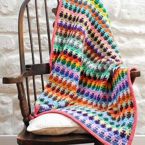 May include: A colorful crocheted blanket with a pink border draped over a wooden rocking chair. The blanket features a geometric pattern in shades of red, orange, yellow, green, blue, purple, and brown.