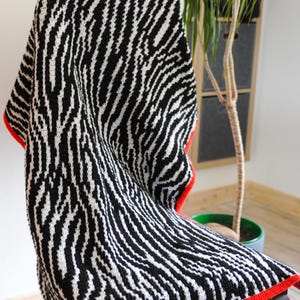 May include: A black and white crocheted blanket with a zebra stripe design, draped over a chair. The blanket features a red border. This blanket is a cozy home decor item.