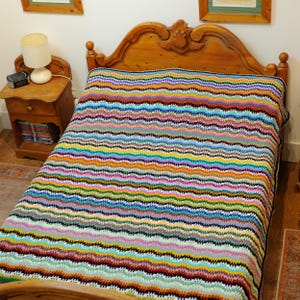 Könnte beinhalten: Eine farbenfrohe, handgehäkelte Decke mit Zickzackmuster. Die Decke zeigt eine breite Palette von Farben, darunter Blau, Grün, Gelb, Rot und Lila. Sie liegt auf einem hölzernen Bettrahmen und unterstreicht ihr lebendiges Design und ihre Textur.