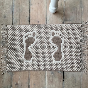 Puede incluir: Una alfombra de baño rectangular, tejida a mano, con un patrón geométrico marrón y crema. La alfombra presenta dos huellas de pies marrones en el centro. Los bordes están acabados con flecos.