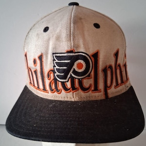 Op de afbeelding: Een beige honkbalpet met een zwarte klep en een zwarte knoop aan de bovenkant. De voorkant is voorzien van het zwart-oranje Philadelphia Flyers-logo in het midden, met het woord Philadelphia geborduurd in oranje met zwarte omlijningen.