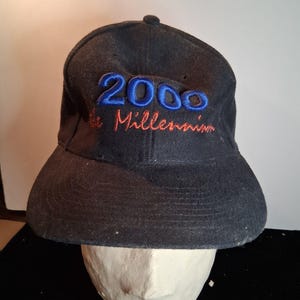 Op de afbeelding: Een zwarte baseballcap met de geborduurde tekst "2000 the Millennium" in blauw en rood. De cap heeft een gebogen klep en een kleine knoop aan de bovenkant. De cap wordt getoond op een mannequinhoofd.
