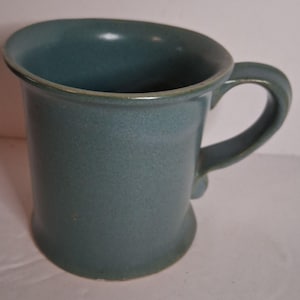 May include: A solid blue-grey ceramic mug with a smooth, matte finish. The mug features a wide, flared rim, a cylindrical body that tapers slightly toward the center, and a sturdy, curved handle attached to the side for a comfortable grip.
