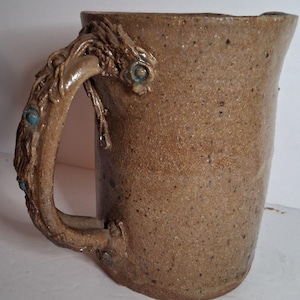 May include: A tan speckled ceramic pitcher with a textured, branch-like handle. The handle features intricate, sculpted details and small, embedded circular turquoise-colored accents. The pitcher has a cylindrical body with a wide, open top.