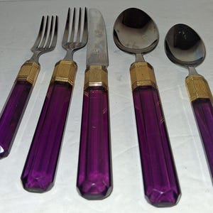 May include: A five-piece flatware set featuring silver-toned metal heads and translucent purple faceted handles. Each piece includes a gold-toned ribbed metal collar connecting the handle to the utensil head, consisting of two forks, a knife, and two spoons.