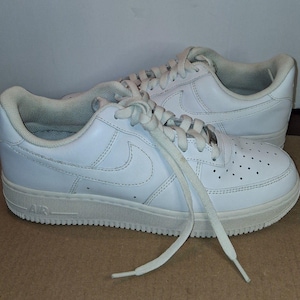 Pode incluir: Um par de tênis de couro branco com o logotipo da Nike na lateral. Os sapatos têm cadarços brancos e uma sola branca com a palavra "AIR" em relevo. Os tênis estão em uma superfície marrom.