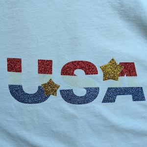 Könnte beinhalten: Ein weißes T-Shirt mit dem Schriftzug USA in einem horizontalen dreifarbigen Muster aus rotem, weißem und blauem Glitzer. Die Buchstaben sind mit zwei goldenen Glitzersternen verziert, einer zwischen U und S und einer über dem Buchstaben A.