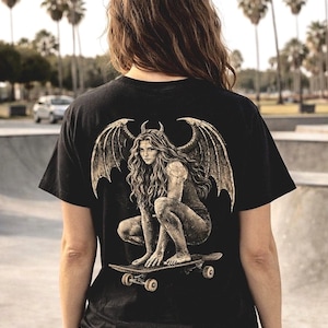 May include: Black t-shirt featuring a detailed graphic of a winged figure with horns, squatting on a skateboard. The illustration is in shades of gray and tan. The background shows a skate park with palm trees.