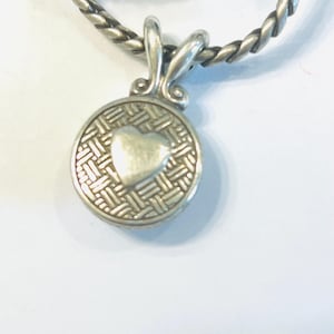 May include: A silver-toned circular pendant featuring a raised smooth heart at the centre, surrounded by a textured basket-weave pattern. The pendant is attached to a thick, twisted silver-toned chain with a polished, metallic finish.