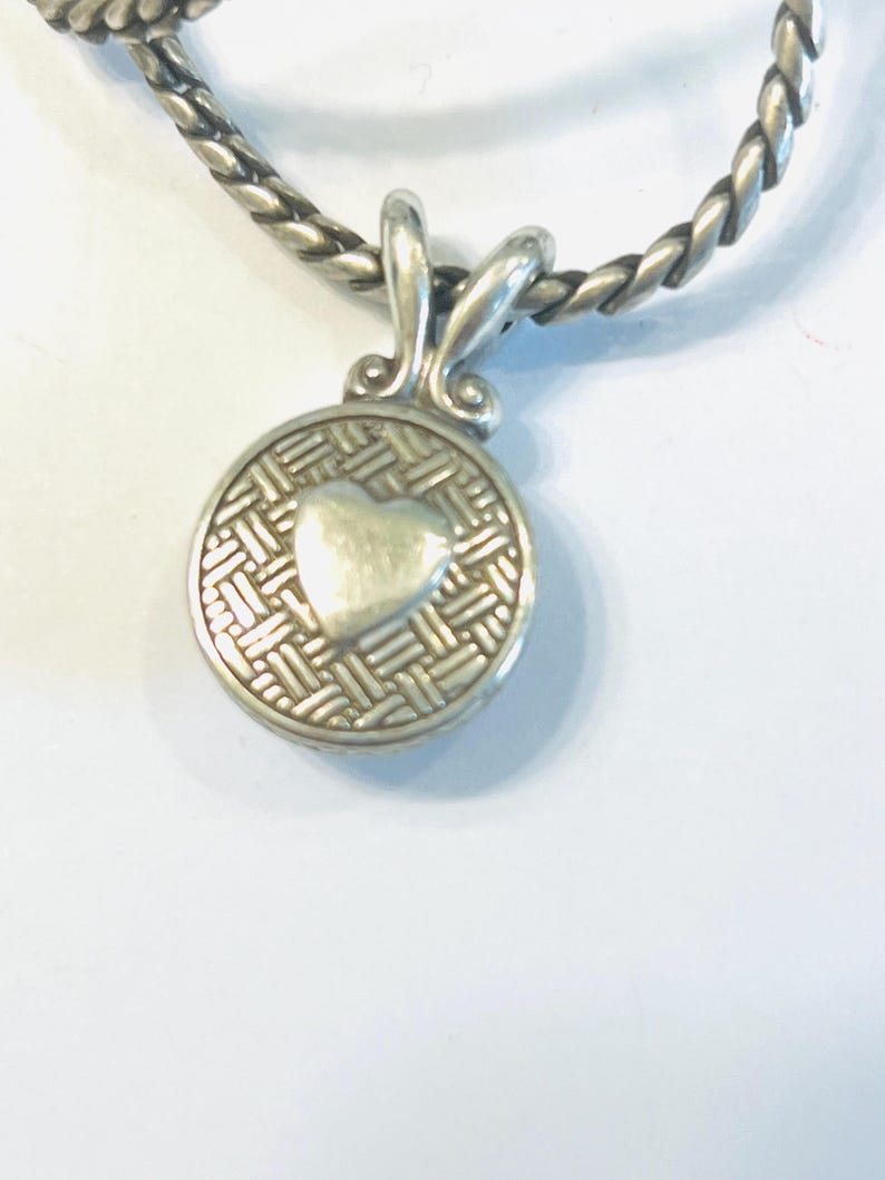 May include: A silver-toned circular pendant featuring a raised smooth heart at the centre, surrounded by a textured basket-weave pattern. The pendant is attached to a thick, twisted silver-toned chain with a polished, metallic finish.