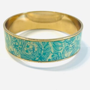 May include: Gold-toned bangle bracelet with a turquoise and cream-colored pattern. The pattern features stars and other shapes. The bracelet has a smooth, polished finish and is suitable for everyday wear.