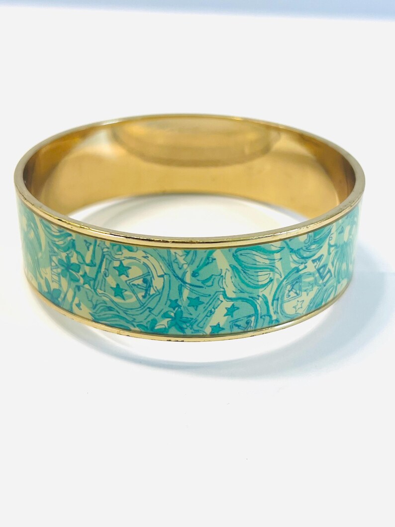 May include: Gold-toned bangle bracelet with a turquoise and cream-colored pattern. The pattern features stars and other shapes. The bracelet has a smooth, polished finish and is suitable for everyday wear.