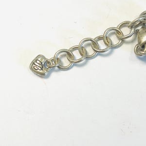 May include: A silver-toned metal chain extender featuring a series of circular links and a small heart-shaped charm at the end. The charm is embossed with the stylised letters TB in a cursive script, providing a decorative finish to the jewellery piece.