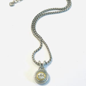 May include: A silver-toned necklace featuring a twisted rope chain and a circular pendant. The pendant has a central clear round gemstone surrounded by a thin gold-toned ring and a textured silver-toned border with a rope-like design.