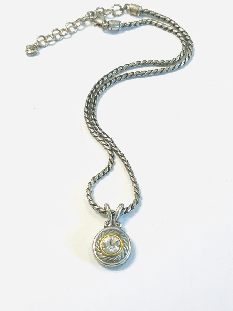 May include: A silver-toned necklace featuring a twisted rope chain and a circular pendant. The pendant has a central clear round gemstone surrounded by a thin gold-toned ring and a textured silver-toned border with a rope-like design.