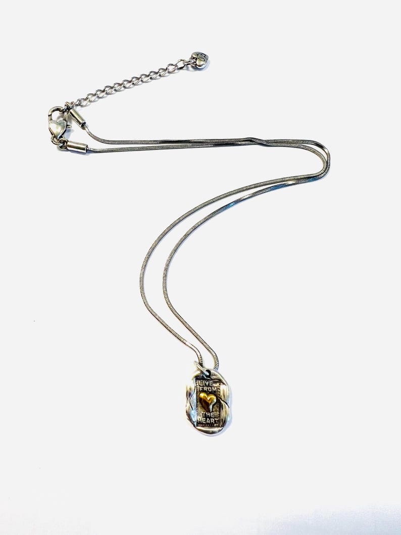 May include: Silver-toned necklace with a pendant featuring a gold-colored heart. The pendant has the words "LIVE FROM THE HEART" engraved on it. The necklace has a delicate chain and a lobster clasp.