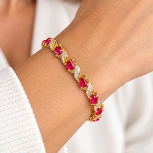 May include: A gold-tone bracelet featuring a series of oval-cut vibrant pinkish-red stones alternating with curved, pavé-set clear stone accents. The metal links are interconnected in a twisted, wave-like pattern throughout the entire piece.