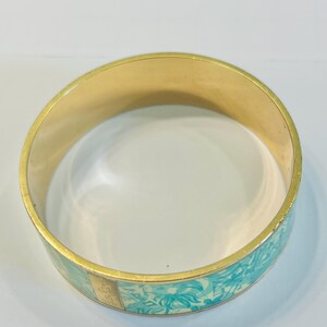 May include: A gold-toned bangle bracelet with a light blue floral pattern. The bracelet has a smooth, polished finish and a rectangular gold accent. The floral design features stylized flowers and leaves in shades of blue and white.
