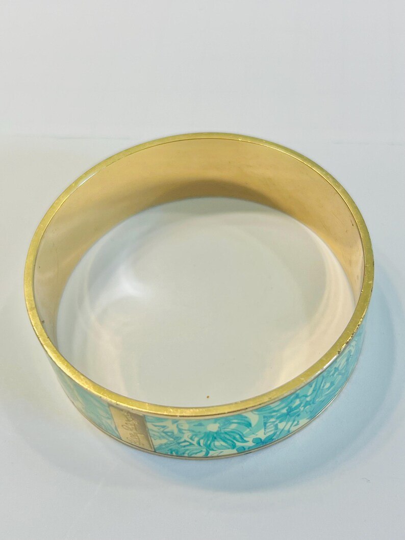 May include: A gold-toned bangle bracelet with a light blue floral pattern. The bracelet has a smooth, polished finish and a rectangular gold accent. The floral design features stylized flowers and leaves in shades of blue and white.