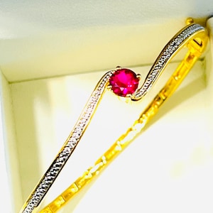 May include: A gold-toned bracelet with a wave design. The bracelet features a vibrant red gemstone at its center and is accented with small, sparkling stones along one side. The jewelry is displayed against a soft, off-white background.