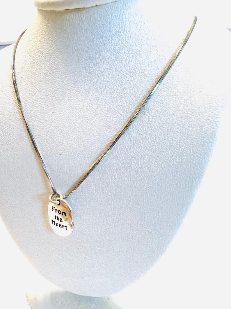May include: A silver-toned necklace with a delicate chain and a small, oval pendant. The pendant is engraved with the words "From the heart" in a simple, elegant font. The necklace is displayed on a white mannequin.