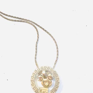 Puede incluir: Collar plateado con un colgante con un dise&ntilde;o de Mickey Mouse. El colgante tiene un marco circular con recortes en forma de coraz&oacute;n y una cabeza central de Mickey Mouse con una gema en forma de coraz&oacute;n. La cadena del collar es de estilo delicado y tejido.