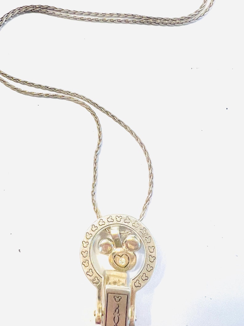 Puede incluir: Collar plateado con un colgante con un dise&ntilde;o de Mickey Mouse. El colgante tiene un marco circular con recortes en forma de coraz&oacute;n y una cabeza central de Mickey Mouse con una gema en forma de coraz&oacute;n. La cadena del collar es de estilo delicado y tejido.