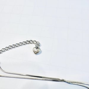 May include: Silver-toned necklace with a heart-shaped charm. The charm features an engraved design. The necklace has a delicate chain and a longer, thinner chain. The background is a white surface.