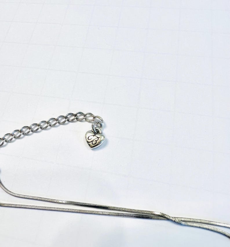 May include: Silver-toned necklace with a heart-shaped charm. The charm features an engraved design. The necklace has a delicate chain and a longer, thinner chain. The background is a white surface.